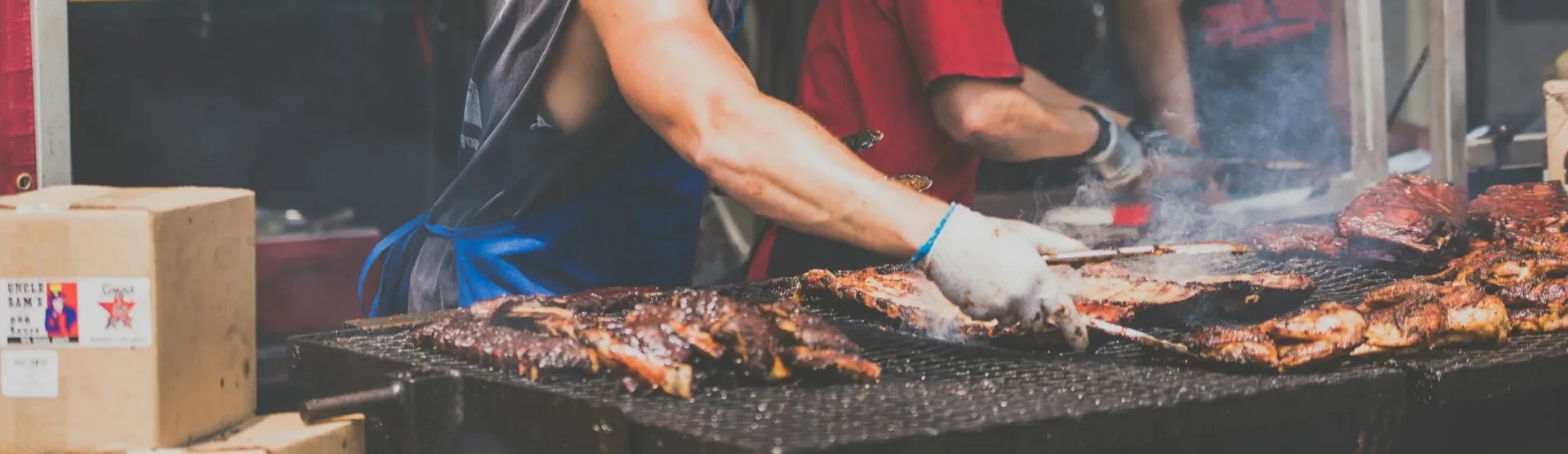 man grilling ribs outside, a mareket. He is wearing a sleeveless shirt and is wearing shades, there is also two people behind him a man and a woman helping out and preparing the meat